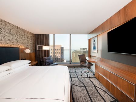 A modern hotel room features a large bed, wall-mounted TV, desk, and chair, with a window view of city buildings and a body of water.
