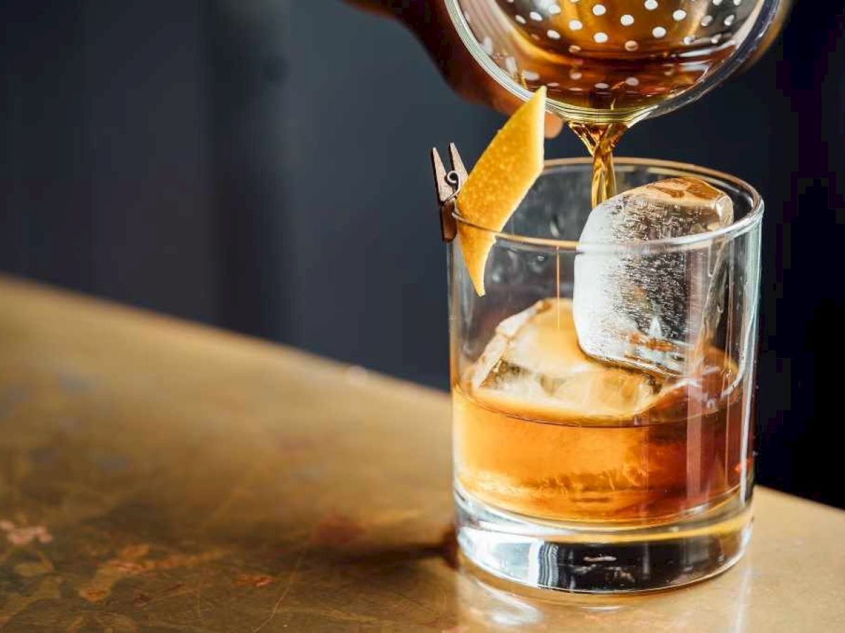A hand pours a drink into a glass with ice and a citrus peel garnish on a wooden surface.