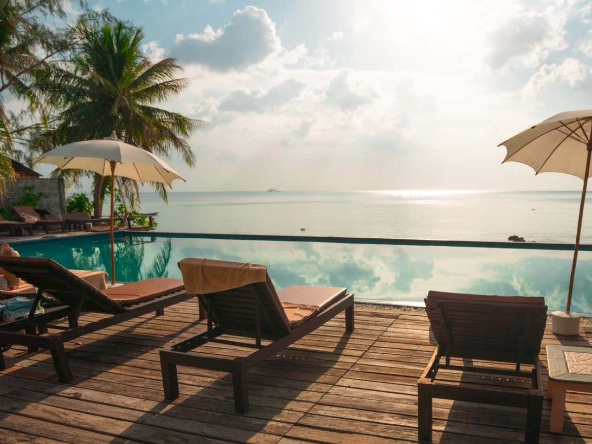 A serene beachside scene with lounge chairs, parasols, and a view of the ocean under a bright sky, surrounded by palm trees.