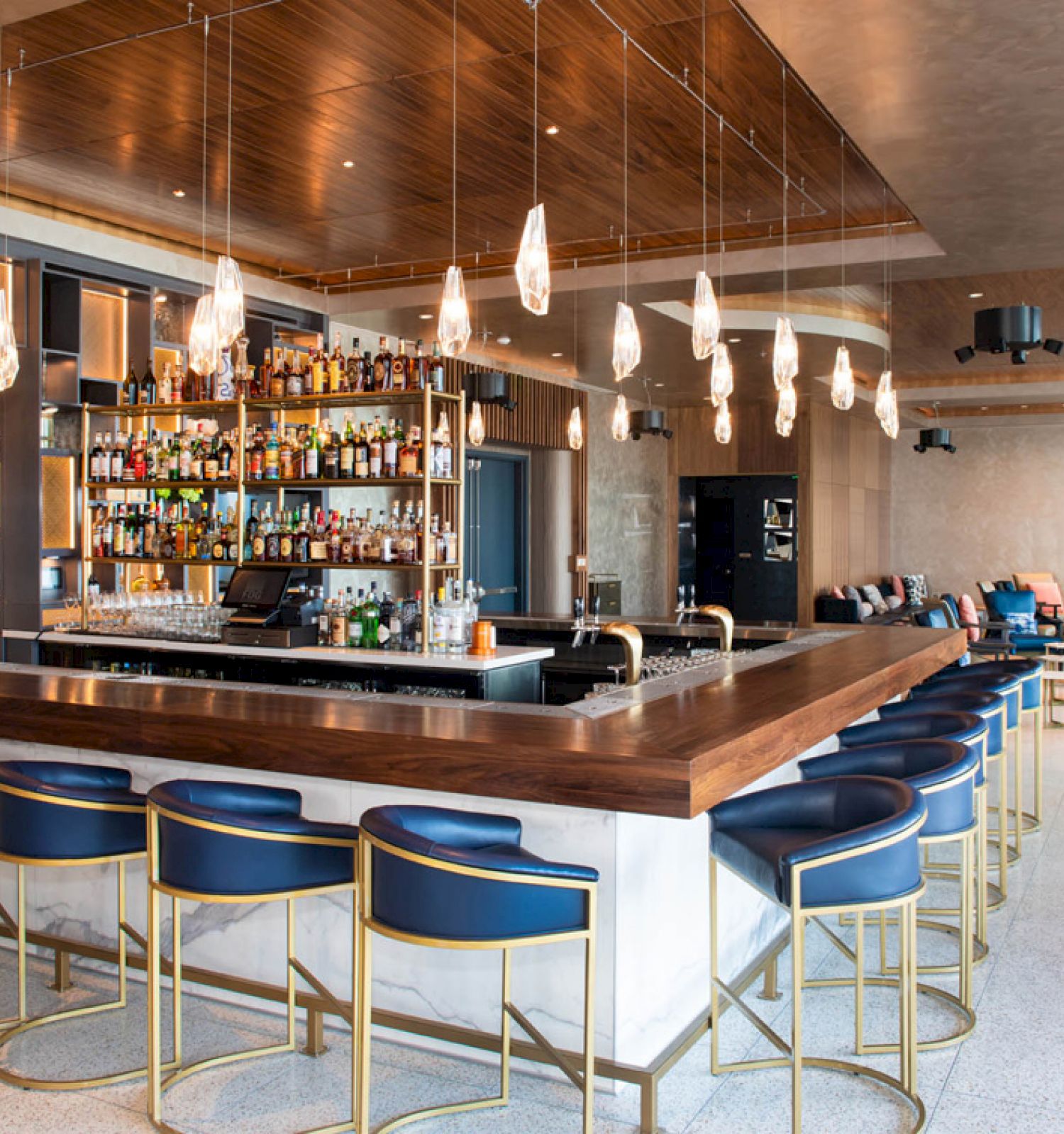A modern bar with suspended lights, a fully stocked shelf, and sleek blue stools surrounding a wooden countertop.