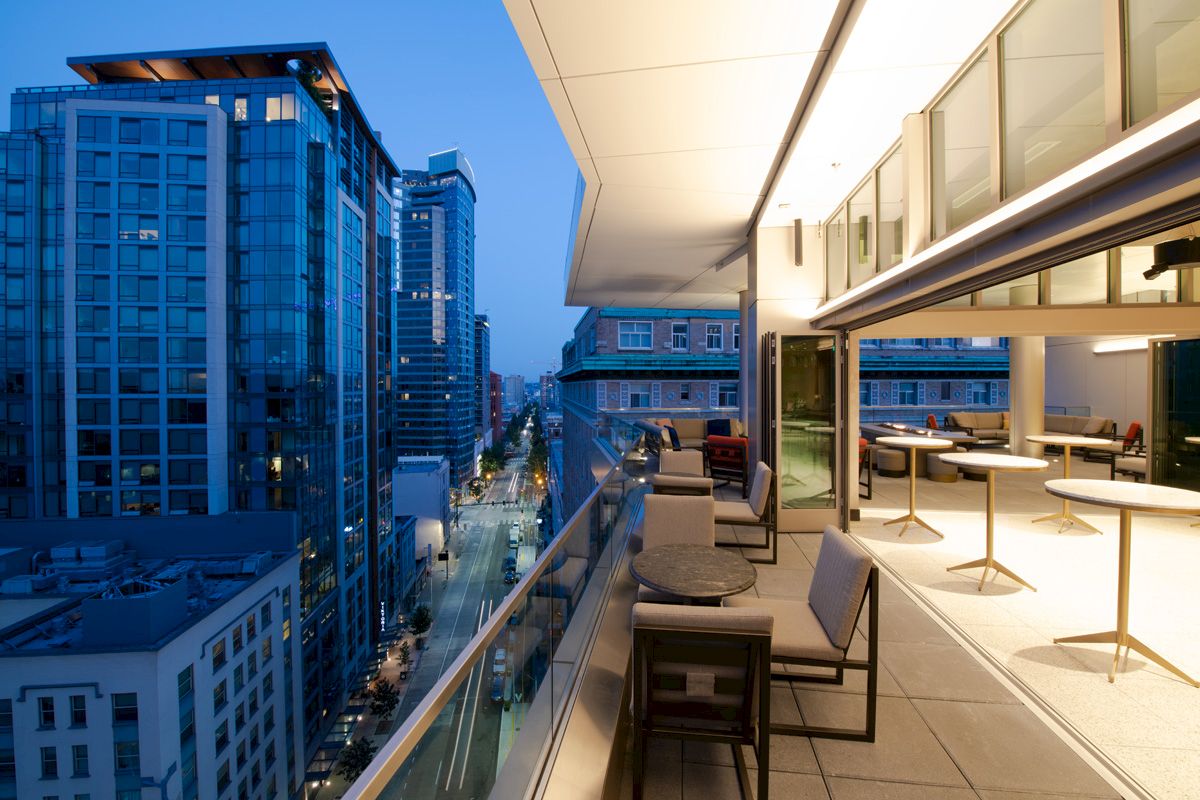 A modern rooftop terrace at dusk overlooks a cityscape with tall buildings, featuring seating areas and a sleek design.