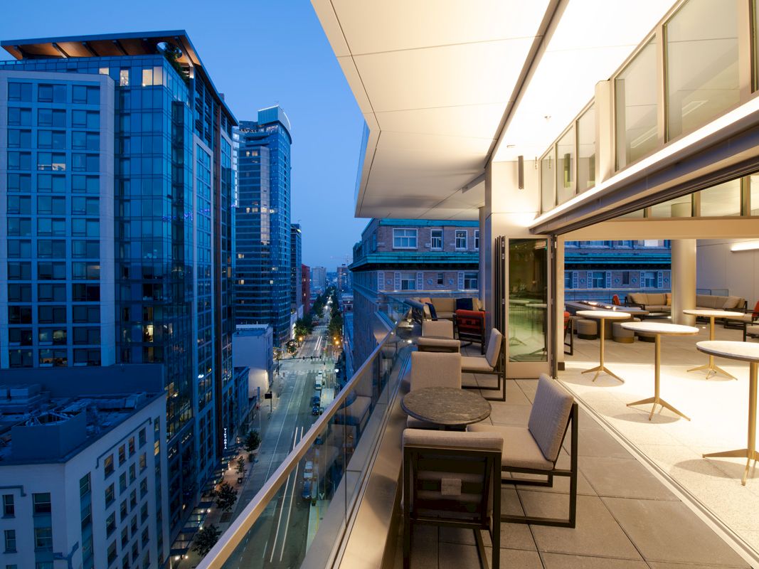A modern rooftop terrace at dusk overlooks a cityscape with tall buildings, featuring seating areas and a sleek design.