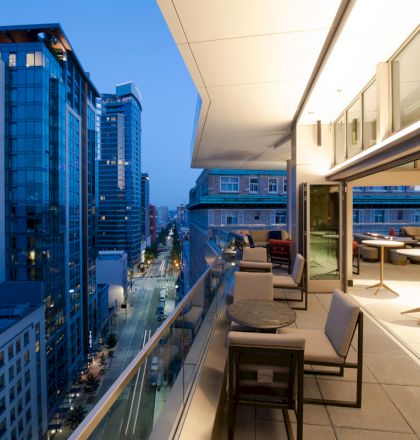 A modern urban balcony at dusk with seating, overlooking a cityscape of tall glass buildings and a lit street.