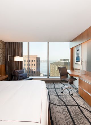 A modern hotel room with a large bed, desk, chair, flat-screen TV, and a window view of buildings outside.