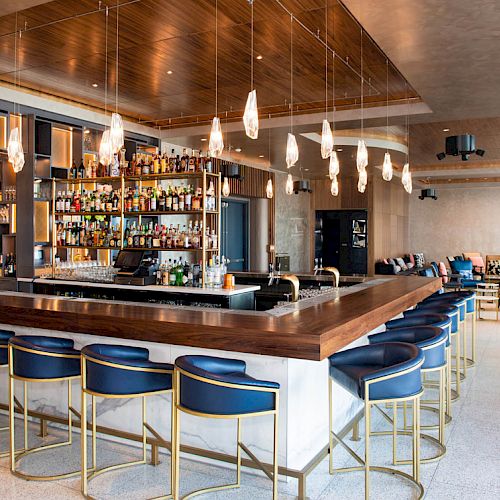 The image shows a modern bar with a large counter, bar stools, hanging lights, and a well-stocked shelf of bottles.