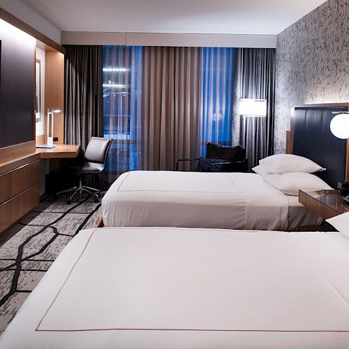 The image shows a modern hotel room with two beds, a wall-mounted TV, a desk, and lamps, featuring a neutral color palette and patterned carpet.