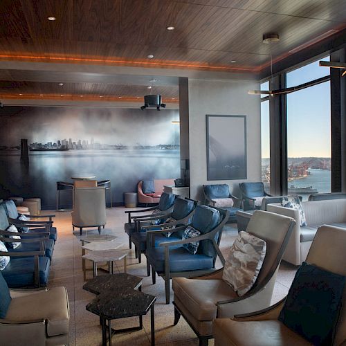 A modern lounge with plush chairs, large windows offering a scenic view, and ambient lighting. A cityscape mural decorates one wall.