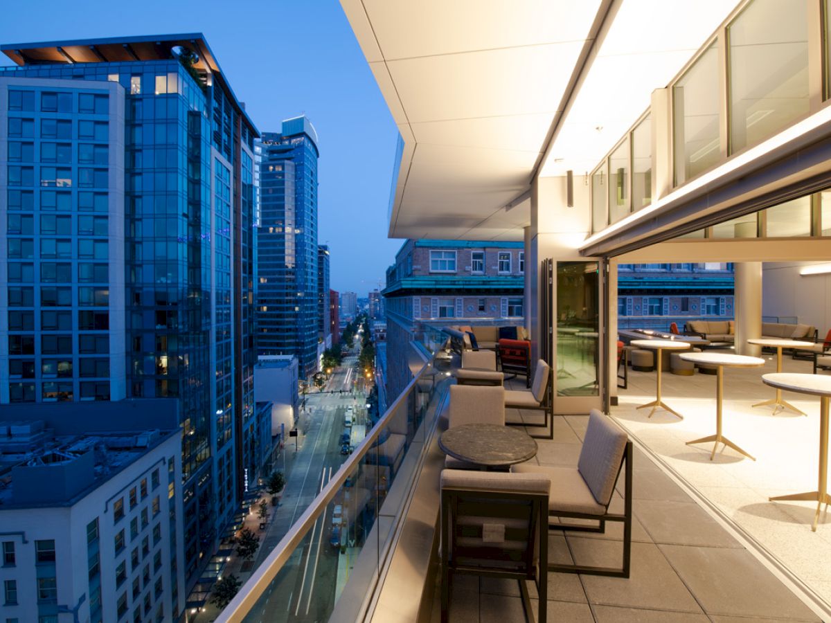 A modern city balcony at twilight shows tables and chairs with a view of tall buildings and a street below.