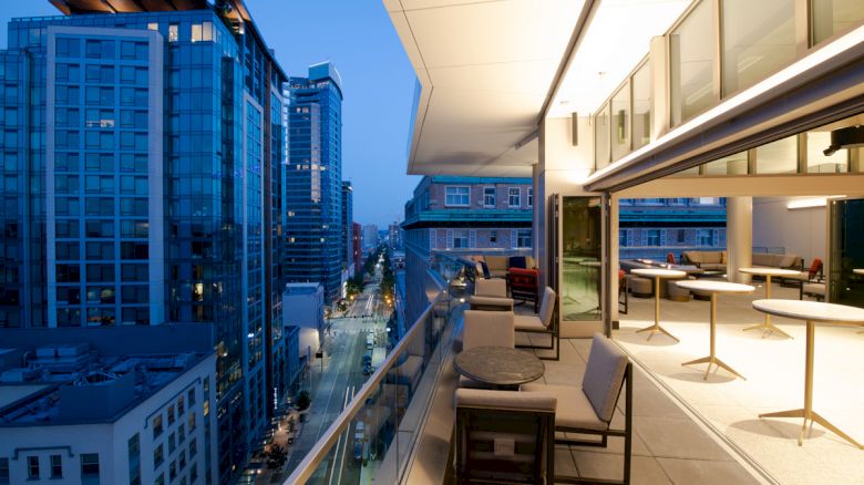 A modern city balcony at twilight shows tables and chairs with a view of tall buildings and a street below.