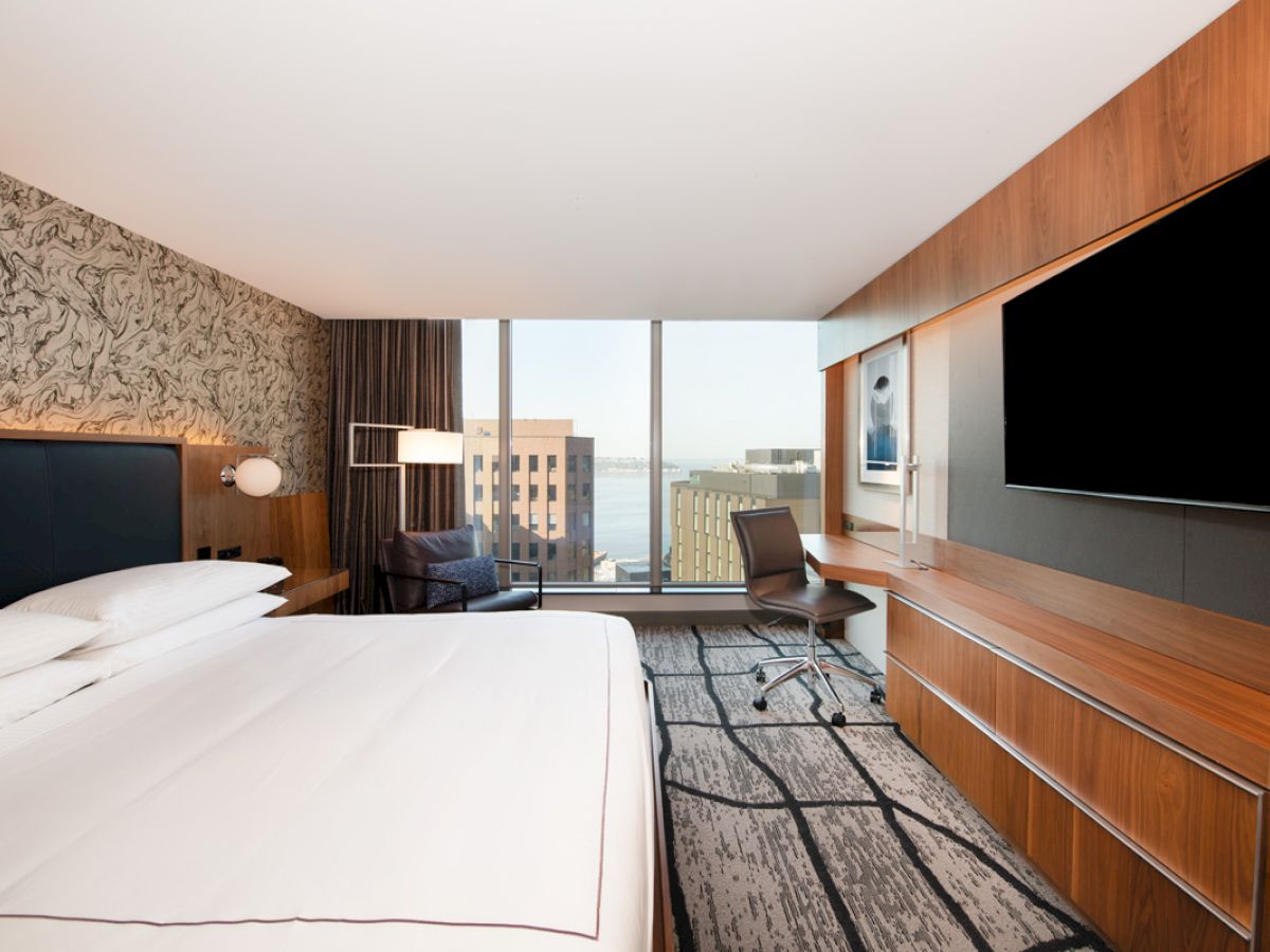 A modern hotel room with a large bed, wall-mounted TV, desk, chair, and a window offering a city view.