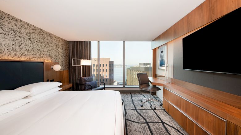 A modern hotel room with a large bed, wall-mounted TV, desk, chair, and a window offering a city view.