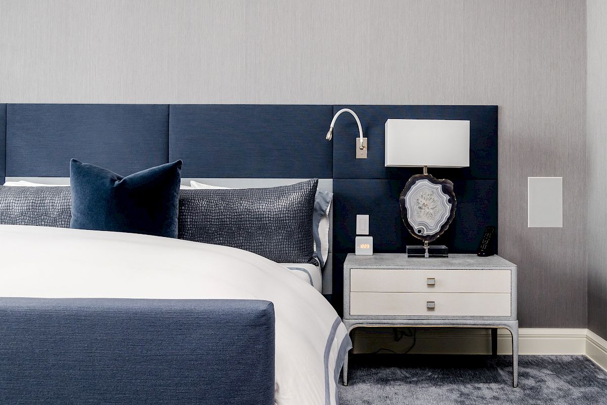 A modern bedroom with a blue headboard, cozy bedding, and a bedside table with a lamp and decor on a soft carpeted floor.