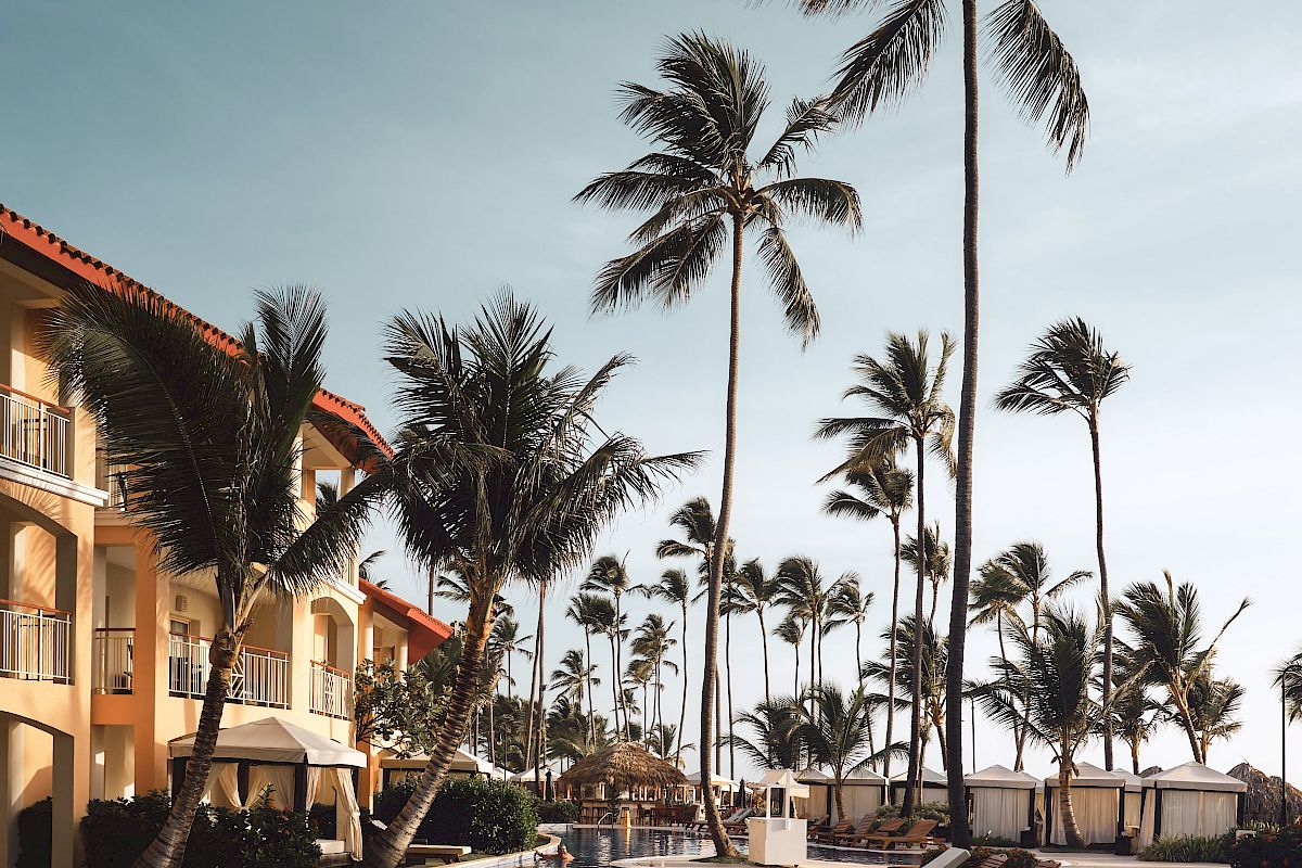 A tropical resort scene featuring tall palm trees, a clear blue sky, and a tranquil pool surrounded by lounge chairs and a building.