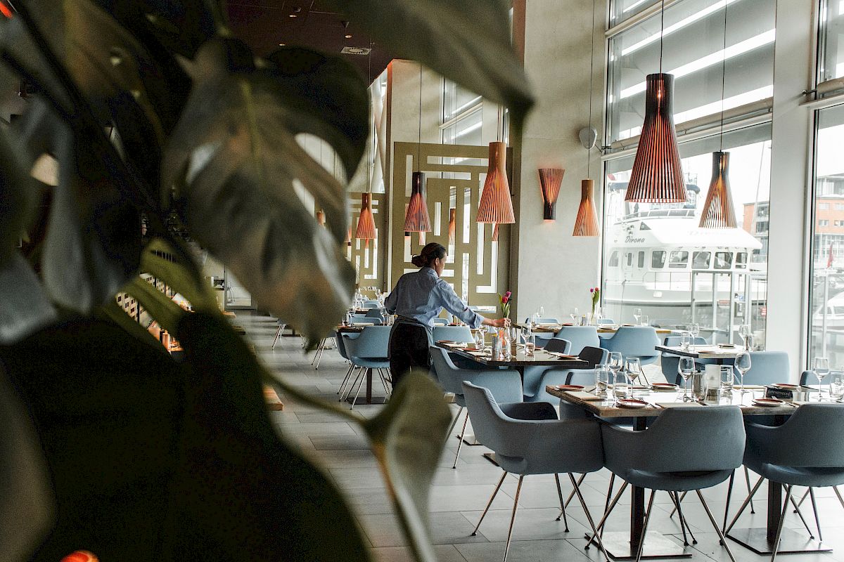 A person is setting tables in a modern restaurant with stylish lighting and large windows.