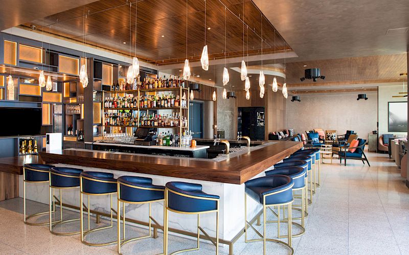 A stylish bar with a wooden countertop, blue bar stools, overhead lights, and a well-stocked shelf of bottles, in a modern lounge setting.