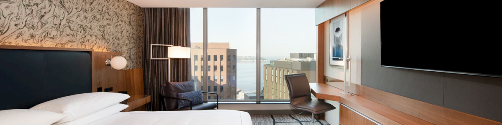 A modern hotel room features a large bed, wall-mounted TV, desk, and city view through a large window, with contemporary decor.